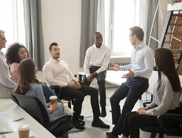 A diverse team of professionals sitting in a modern office listening to a male leader explain strategic business goals and executive coaching benefits.